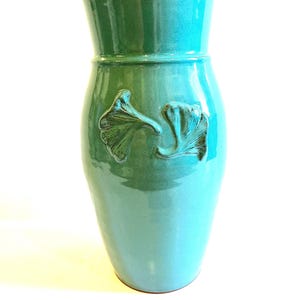 May include: A tall, turquoise ceramic vase with a glossy finish. The vase features two raised ginkgo leaf designs and a flared top. The color transitions from a darker teal at the top to a lighter turquoise at the base. A decorative piece for the home.