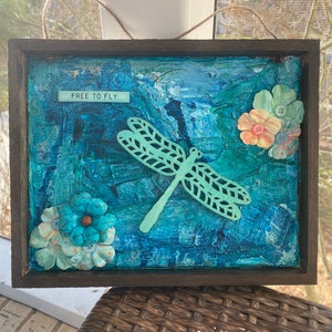 Peut inclure: Une œuvre d'art encadré en techniques mixtes représentant une libellule bleue et verte avec les mots "Free to Fly" sur un fond bleu avec des fleurs en papier.