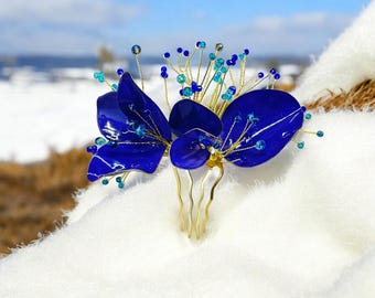 May include: A vibrant blue hair comb featuring a floral design. The comb has three large, glossy blue petals and smaller blue and turquoise beads. The comb's base is gold-colored, with several prongs for securing it in the hair. The background is a snowy landscape.