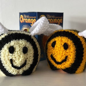 May include: Two crocheted bee toys with black and white stripes and yellow and black stripes. The bees have white wings and smiling faces. The bees are sitting in front of two boxes of Terry's Chocolate Orange.