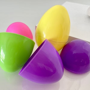 May include: A collection of colorful plastic Easter eggs in various shades. The image features a bright yellow egg, a pink egg, a green egg, and a purple egg. The eggs are open and closed, suggesting they are for hiding treats or small toys.