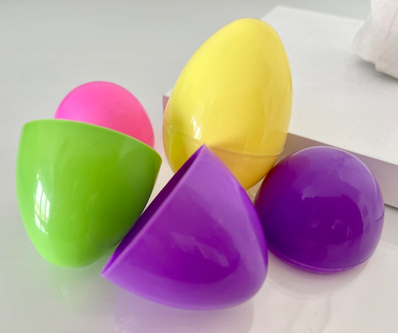 May include: A collection of colorful plastic Easter eggs in various shades. The image features a bright yellow egg, a pink egg, a green egg, and a purple egg. The eggs are open and closed, suggesting they are for hiding treats or small toys.