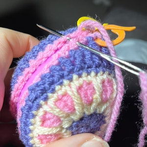 May include: Close-up of a crochet project in progress. The project features a colorful design with pink, purple, and cream yarn. A needle and stitch marker are visible, highlighting the handmade nature of the item.