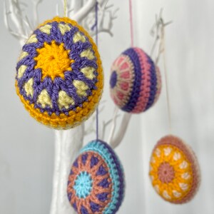 May include: Four colorful, crocheted Easter egg ornaments hanging from a white decorative tree. The eggs feature intricate patterns in shades of yellow, purple, blue, pink, and brown. Each egg is suspended by a thin string.