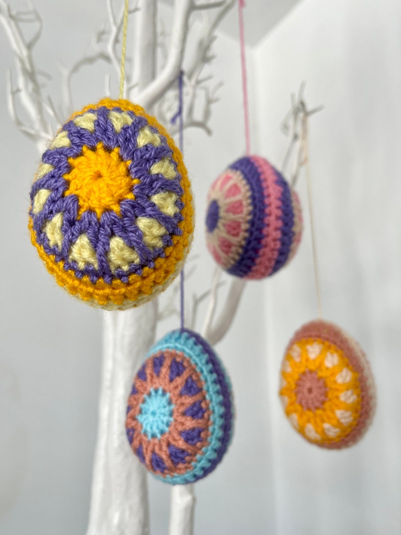 May include: Four colorful, crocheted Easter egg ornaments hanging from a white decorative tree. The eggs feature intricate patterns in shades of yellow, purple, blue, pink, and brown. Each egg is suspended by a thin string.