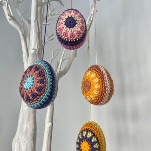 May include: Four crocheted Easter egg ornaments in various colors, hanging from a white decorative tree. The eggs feature intricate patterns in shades of blue, purple, pink, yellow, and brown. Each egg is suspended by a thin string, creating a festive display.