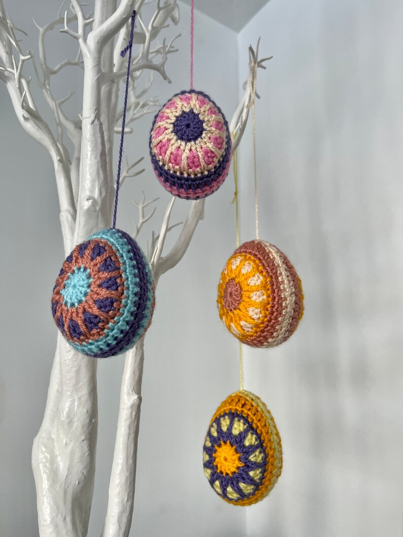 May include: Four crocheted Easter egg ornaments in various colors, hanging from a white decorative tree. The eggs feature intricate patterns in shades of blue, purple, pink, yellow, and brown. Each egg is suspended by a thin string, creating a festive display.
