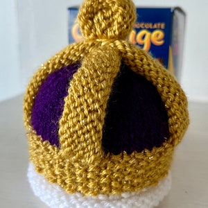 Puede incluir: Una corona de ganchillo con la parte superior morada, ribete dorado y base blanca. La corona es un artículo hecho a mano.
