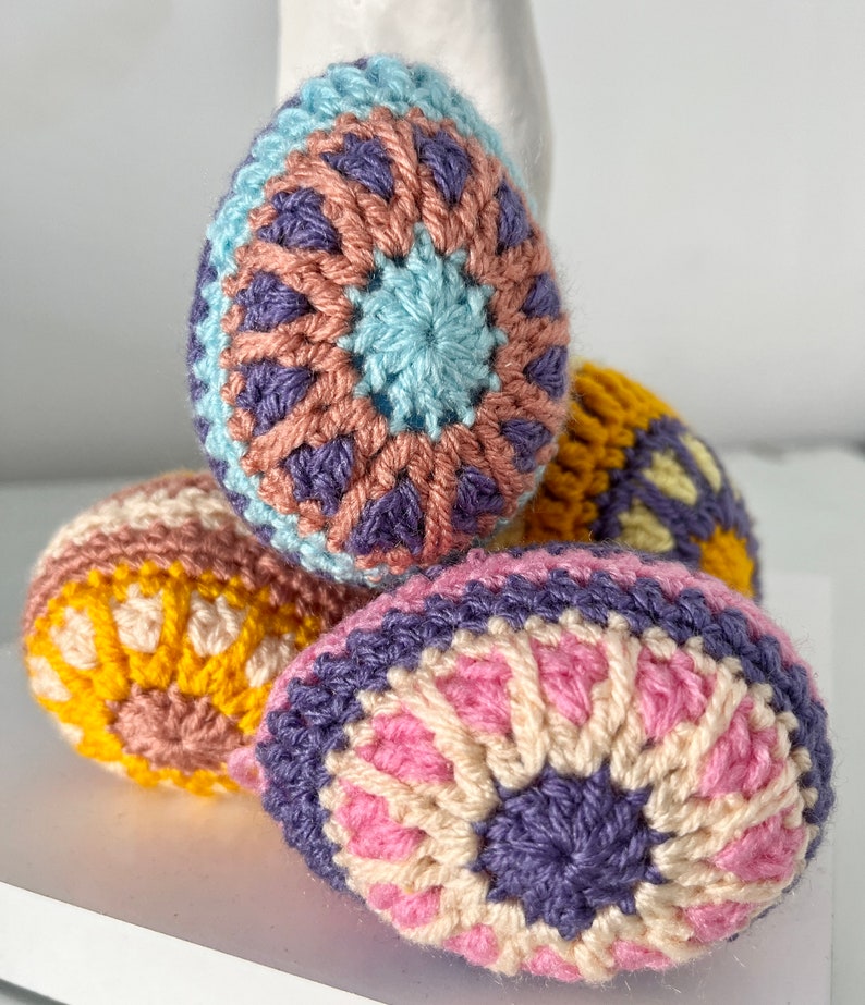 May include: Four crocheted Easter eggs in various colors. The eggs feature intricate patterns in shades of blue, pink, purple, yellow, and cream. The eggs are arranged on a white surface, showcasing the handmade details and vibrant colors.