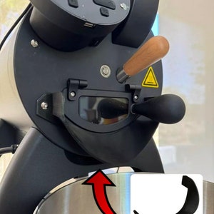 Könnte beinhalten: Ein schwarzer Kaffeeröster mit einem Auffangbehälter aus Edelstahl für die Bohnen. Der Röster hat einen Holzgriff und eine schwarze Kunststoff-Bohnenrinne. Die Bohnenrinne ist mit einer schwarzen Metallhalterung am Röster befestigt. Die Halterung ist separat im Bild dargestellt.