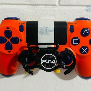 Op de afbeelding: Oranje en zwarte PlayStation 4-controller met een zwart accessoire. De controller heeft blauwe knoppen en een D-pad. Het accessoire toont het PS4-logo in wit. De controller is op een wit oppervlak gemonteerd.