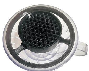 Könnte beinhalten: Ein transparenter Kaffeezubereiter mit einem schwarzen Filter im Wabenmuster. Der Zubereiter hat einen Griff und einen schwarzen Ring am oberen Rand. Das Design ermöglicht einen kontrollierten und gleichmäßigen Kaffeebrühvorgang.