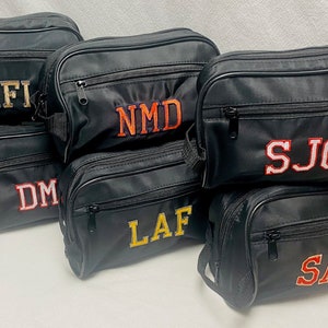 May include: Six black zippered travel bags with different embroidered initials in various colors. The initials are MFI, NMD, SJG, DM, LAF, and SAR.
