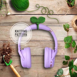 May include: Purple wireless headphones with a green crocheted leaf design on the headband. The headphones are on a wooden surface with gardening supplies and plants.