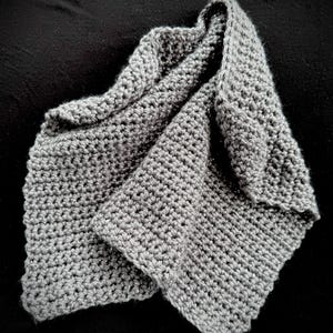 May include: A folded, gray crocheted scarf. The scarf is made of a soft, textured yarn and has a simple, yet elegant design. The scarf is folded over itself, creating a layered effect. The color is a neutral gray, suitable for various outfits.