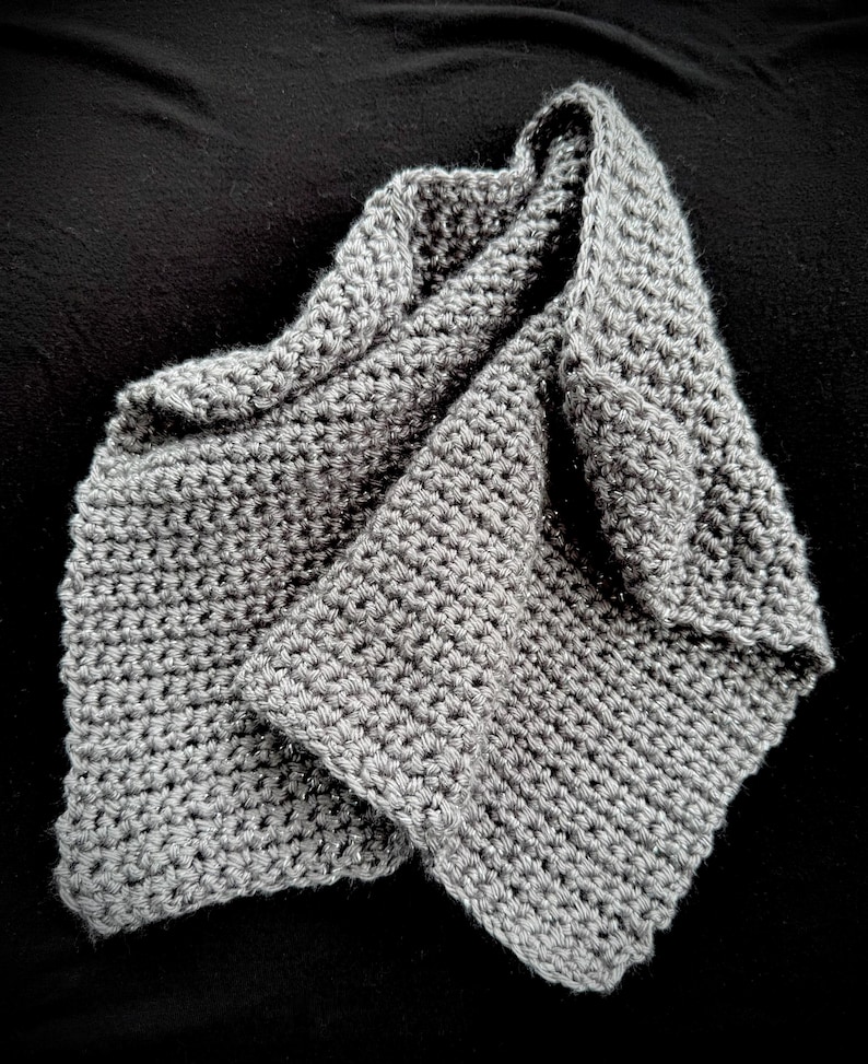 May include: A folded, gray crocheted scarf. The scarf is made of a soft, textured yarn and has a simple, yet elegant design. The scarf is folded over itself, creating a layered effect. The color is a neutral gray, suitable for various outfits.