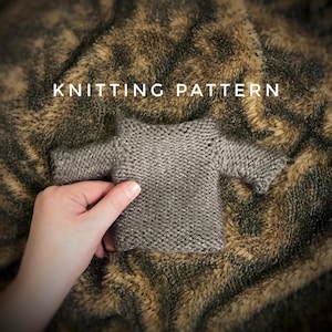 May include: Knitting pattern for a gray knitted sweater with short sleeves. The sweater is lying on a brown and green faux fur background.