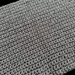 May include: Close-up of a gray, hand-crocheted scarf. The scarf has a textured appearance with a tight stitch pattern. The yarn has a subtle metallic sheen, adding a touch of sparkle. The scarf is displayed against a dark background.