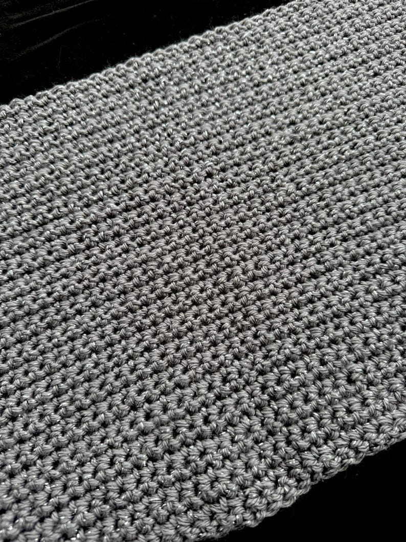 May include: Close-up of a gray, hand-crocheted scarf. The scarf has a textured appearance with a tight stitch pattern. The yarn has a subtle metallic sheen, adding a touch of sparkle. The scarf is displayed against a dark background.