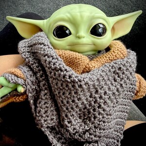 May include: A green-skinned doll with large black eyes and pointed ears is wrapped in a gray and brown crocheted blanket. The doll is a character from a popular science fiction franchise.