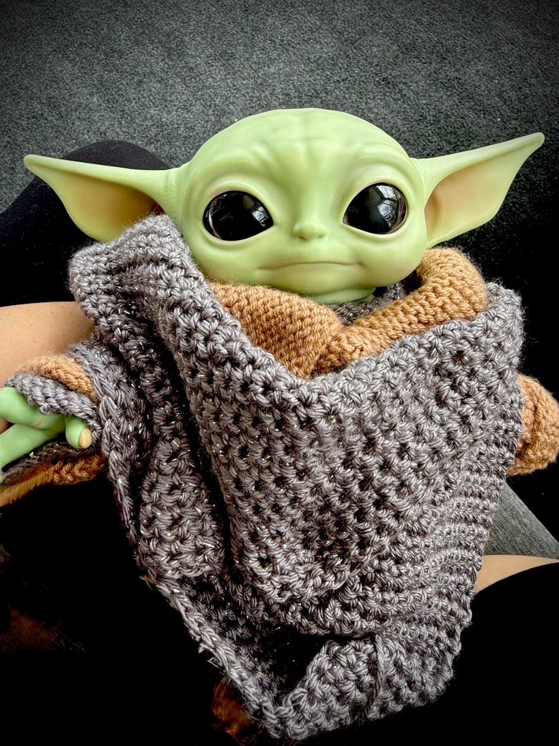 May include: A green-skinned doll with large black eyes and pointed ears is wrapped in a gray and brown crocheted blanket. The doll is a character from a popular science fiction franchise.