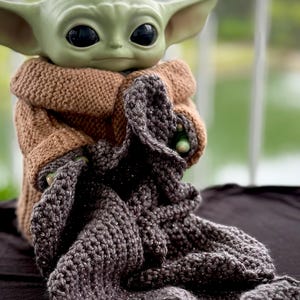 May include: A green-skinned Grogu figure, also known as Baby Yoda, is wrapped in a brown knitted coat and a gray crocheted blanket. The figure has large black eyes and pointed ears. The blanket has a textured appearance.