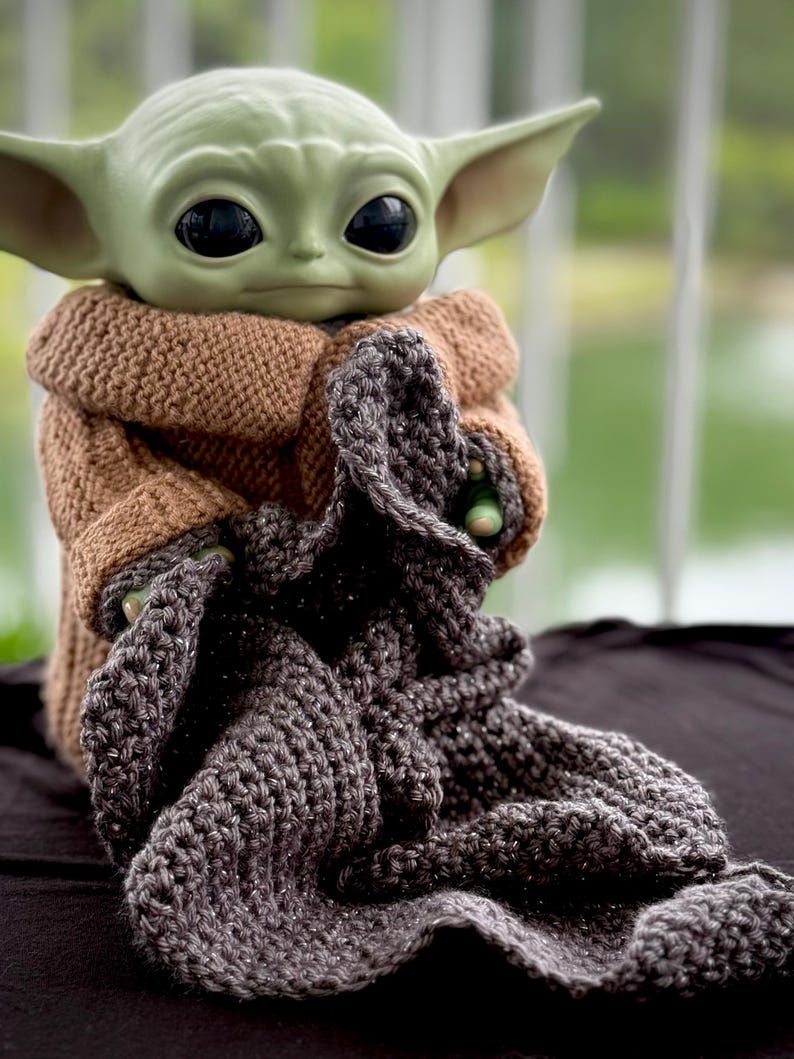 May include: A green-skinned Grogu figure, also known as Baby Yoda, is wrapped in a brown knitted coat and a gray crocheted blanket. The figure has large black eyes and pointed ears. The blanket has a textured appearance.