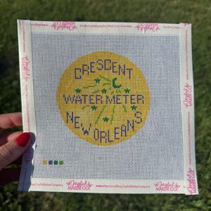 May include: A square needlepoint canvas featuring a yellow circle with the purple text CRESCENT WATER METER NEW ORLEANS and green star and crescent moon accents. The white canvas has a grid pattern and branded tape borders on all four sides.