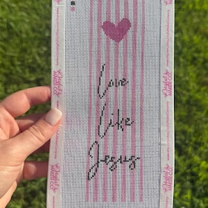 May include: A rectangular needlepoint canvas featuring a vertical pink and white striped pattern. A solid pink heart is centered near the top. Below the heart, the black text reads Love Like Jesus. The edges are branded with Joyful Maker Co.