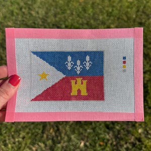 May include: A needlepoint canvas featuring a flag design with blue, red, and white sections. The flag includes a yellow star, fleur-de-lis, and a yellow "H" symbol. The canvas is framed with a pink border.