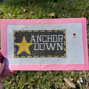 May include: A needlepoint canvas with a pink border and a black center featuring the words "ANCHOR DOWN" and a yellow star. The canvas is rectangular and appears to be hand-stitched.