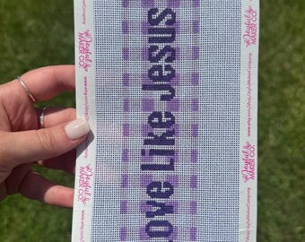 May include: A rectangular needlepoint canvas featuring a purple and white checkered pattern with the text Love Like Jesus printed in dark purple. The edges are bordered by white tape printed with the Joyful Maker Co. logo and URL.