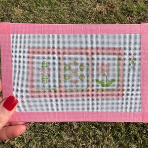 May include: A pink needlepoint canvas with a floral design. The canvas features three squares with green and pink flowers. The canvas is framed with a pink border. The background is white.
