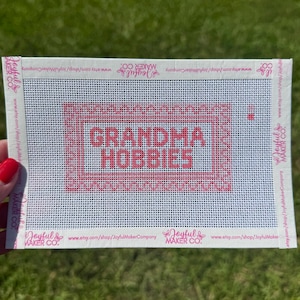 May include: A rectangular white needlepoint canvas featuring a pink stitched border and the text "GRANDMA HOBBIES" in pink. The edges are bound with white tape printed with the text "Joyful Maker Co." and the shop URL in pink ink.