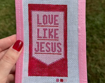 May include: A rectangular needlepoint canvas featuring a pink fabric border and a central design with the text LOVE LIKE JESUS in dark red and pink thread. The background is a white grid mesh with small color swatches at the bottom.