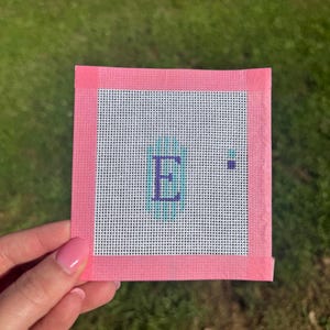 May include: A square needlepoint canvas with a pink border. The canvas features the letter "E" in a teal and purple color scheme. The background is white with a grid pattern.