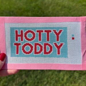 May include: A pink needlepoint with the words "HOTTY TODDY" in red letters on a teal background. The needlepoint is framed in pink and is set against a green grassy background.