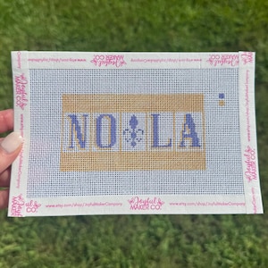 May include: A rectangular needlepoint canvas featuring a gold background with the blue text NO LA and a central blue fleur-de-lis symbol. The white border is printed with the text Joyful Maker Co. and the shop URL repeatedly.
