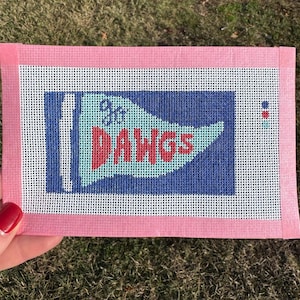 May include: Needlepoint canvas with a pink border. The canvas features a blue flag design with the words "Go Dawgs" in red and light blue. A small red dot is in the upper right corner. The canvas is held by a person.