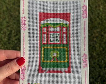 May include: A rectangular needlepoint canvas featuring a green and red streetcar design with the text NOLA in the center window and a wreath on the front panel. The canvas is bordered by white tape printed with the Joyful Maker Co. logo.
