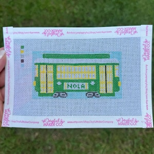 May include: A needlepoint canvas featuring a green streetcar with yellow windows and the text NOLA printed on the side. The canvas includes a color key on the left side and white binding tape along the edges with the text Joyful Maker Company.