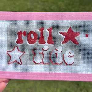May include: A pink needlepoint canvas with the words "roll tide" stitched in red and white. The design includes two stars and an exclamation point. The canvas is framed with a pink border.