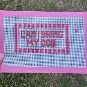 May include: A pink needlepoint canvas with the text "CAN I BRING MY DOG" in red. The canvas has a white grid background and a pink border. The canvas is rectangular.