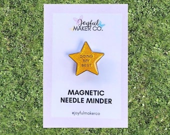 Op de afbeelding: Een gouden, stervormige magnetische naaldenhouder met de tekst "DOING MY BEST" op een witte kaart. De kaart bevat ook de tekst "MAGNETIC NEEDLE MINDER" en de naam van de maker. De achtergrond is groen gras.