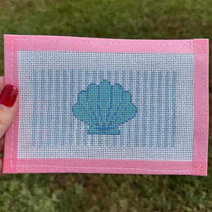May include: A pink needlepoint canvas with a blue seashell design. The canvas has a light blue and white striped background. The seashell is a darker shade of blue. The canvas is held by a hand with red nail polish.