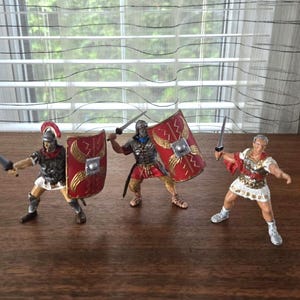 May include: Three miniature Roman soldier figurines standing in a row. The two figures on the left wear dark armor, red crested helmets, and carry rectangular red shields with gold wing designs and swords. The third figure wears white armor.