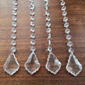 May include: Four clear crystal chandelier pendants with faceted beads and teardrop-shaped drops. The pendants are strung on metal wires and are displayed against a dark wood surface. These decorative elements are suitable for lighting fixtures.
