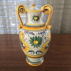 May include: A decorative ceramic vase with two ornate handles. The vase features a cream-coloured body with yellow, blue, and green accents. The design includes floral and mythical creature motifs, with a yellow rim and base. The vase is approximately 25 cm tall.