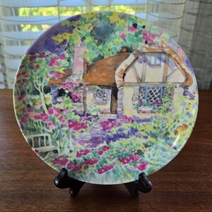 May include: A ceramic plate with a watercolor painting of a cottage with a thatched roof surrounded by a garden with pink flowers. The plate is on a black stand.