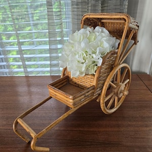 Può includere: Un piccolo risciò decorativo in vimini con un arrangiamento di fiori bianchi all'interno. Il risciò ha una ruota in legno marrone e una carrozzeria in vimini marrone.